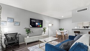 En moderne stue med et fladskærms-tv på væggen, et spiseområde med et hvidt bord og stole, samt en blå sofa med dekorative puder.
