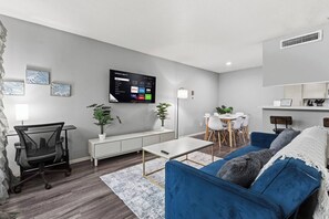 A modern living room with a flat-screen TV mounted on the wall, a dining area with a white table and chairs, and a blue sofa with decorative pillows. - Cozy 1BR in a convenient Austin location. (Austin)