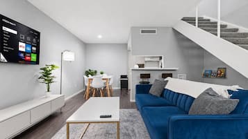 Una sala moderna con un sofá azul, una mesa de comedor y un televisor montado en la pared que muestra servicios de transmisión.