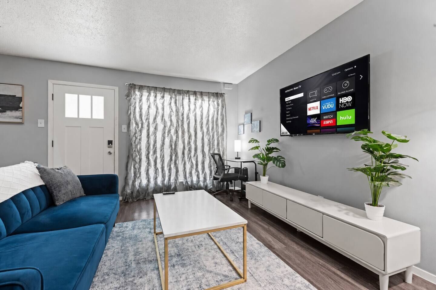 A modern living room with a blue sofa, a white coffee table, a TV mounted on the wall displaying various streaming services, and two potted plants.