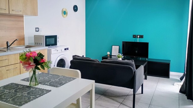 A modern living space with a kitchen area, a washing machine, a dining table with a vase of flowers, and a black sofa. - Appartement Tout Equipe a Lens Avec Parking Gratuit Proche du Louvre Lens (Lens)