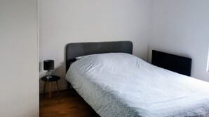 A small, single-bed room with a grey headboard, a bedside table with a lamp, and a plain wall. - Appartement Tout Equipe a Lens Avec Parking Gratuit Proche du Louvre Lens (Lens)