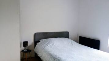 A small, single-bed room with a grey headboard, a bedside table with a lamp, and a plain wall.