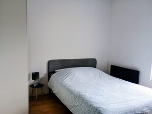 Ein kleines Zimmer mit einem Einzelbett, grauem Kopfteil, Nachttisch mit Lampe und schlichter Wand.