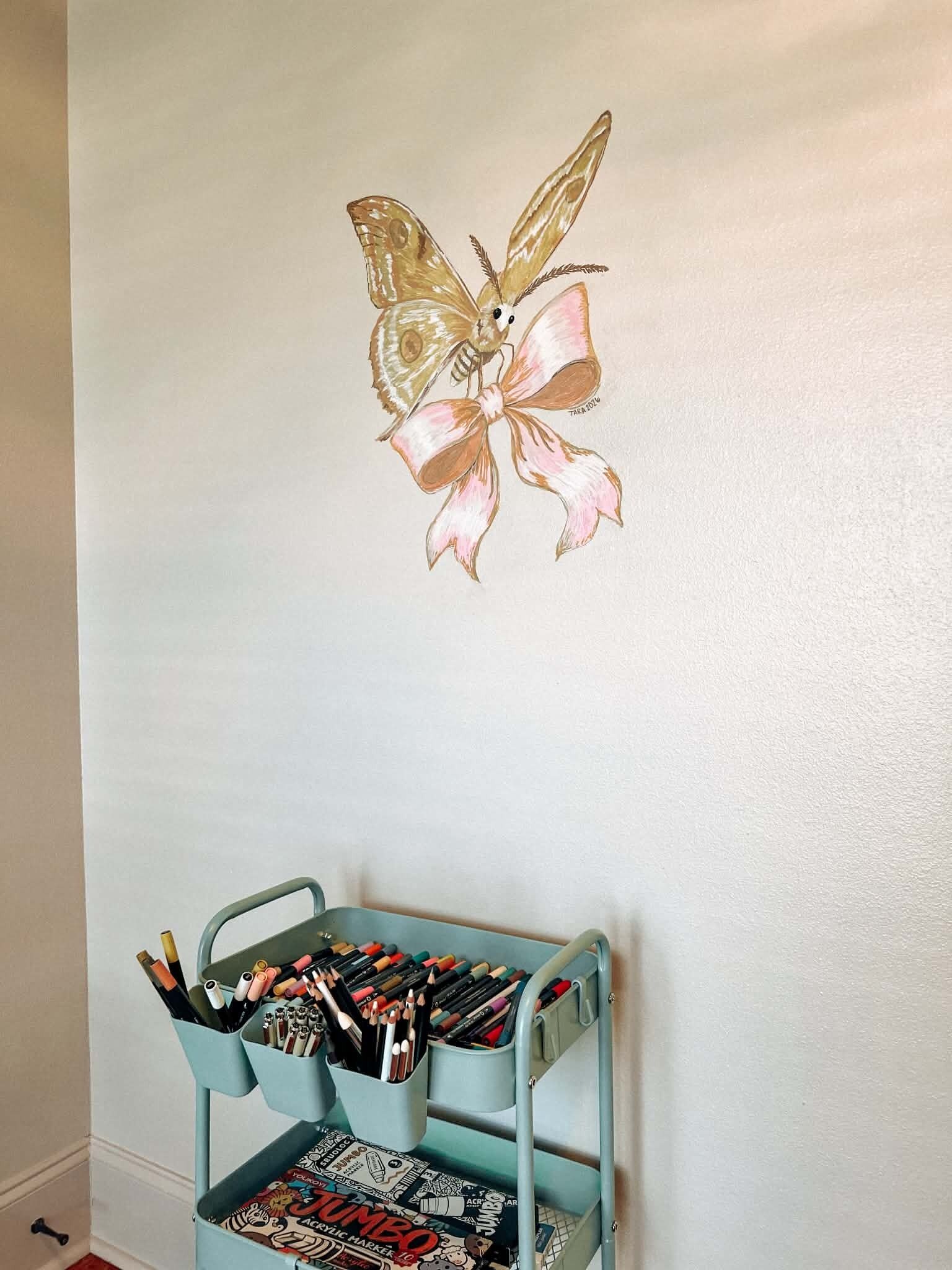 Un mur orné d’une illustration de papillon et un chariot en métal contenant du matériel de dessin.