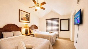 Une chambre d’hôtel avec deux lits, un ventilateur de plafond, une télévision, un tableau encadré et une fenêtre avec des stores.