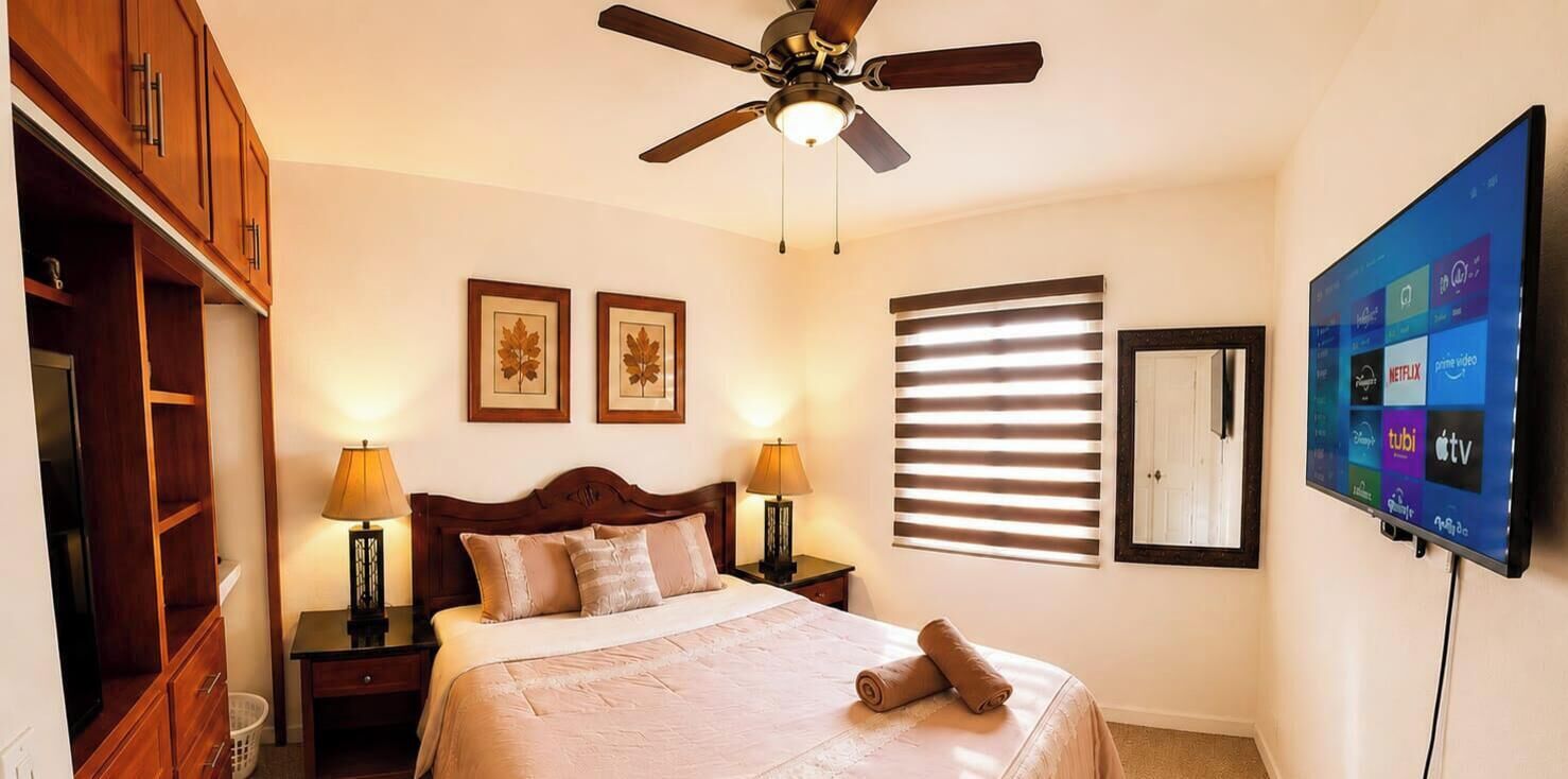 A bedroom with a bed, two bedside lamps, a ceiling fan, a TV mounted on the wall, and a window with blinds.