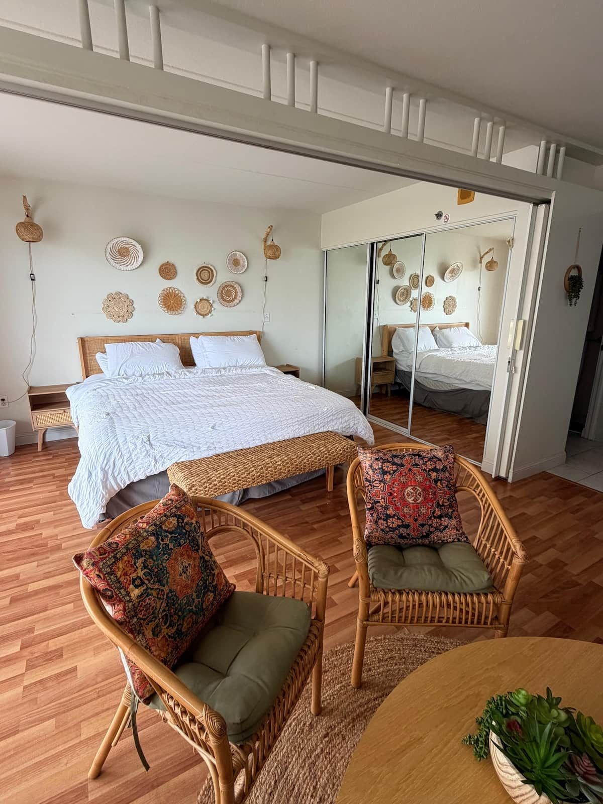 A room with a large bed, two wicker chairs with cushions, a small table with a plant, and a mirror reflecting the bed.