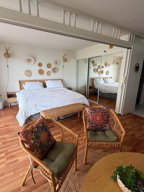 Een kamer met een groot bed, twee rieten fauteuils met kussens, een tafeltje met een plant en een spiegel die het bed weerspiegelt.