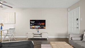 Una sala de estar con una televisión de pantalla plana mostrando 'Stranger Things', un sofá blanco, una mesa de centro de madera y un comedor con una mesa y sillas blancas.