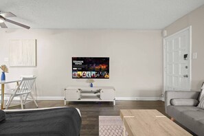 A living room with a flat-screen TV displaying 'Stranger Things', a white sofa, a wooden coffee table, and a dining area with a white table and chairs. - Austin 2BR Home with 3 Beds & Great Location (Austin)