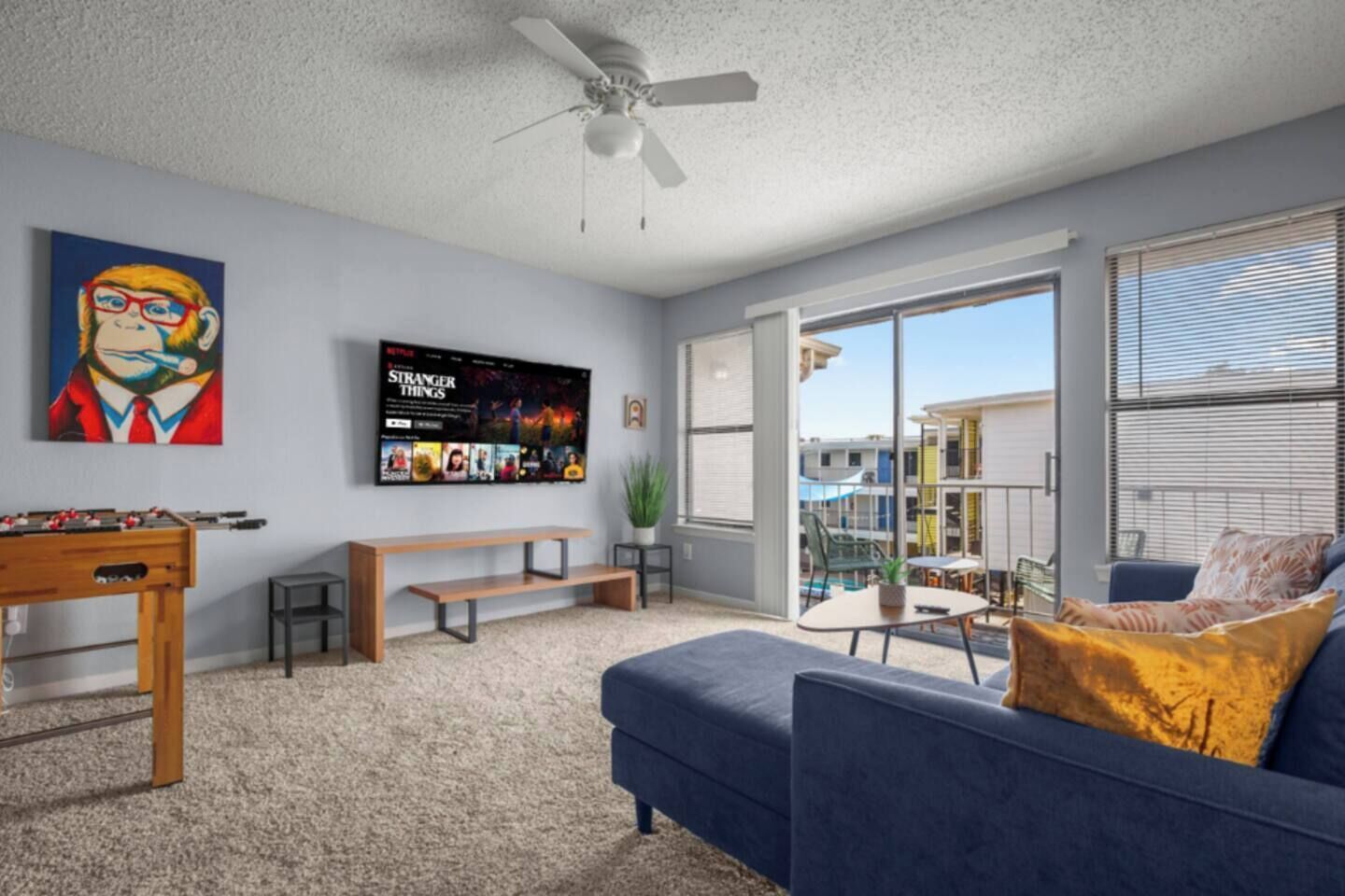 A living room with a sofa, a coffee table, a TV mounted on the wall, a ceiling fan, and a balcony with a view of buildings.