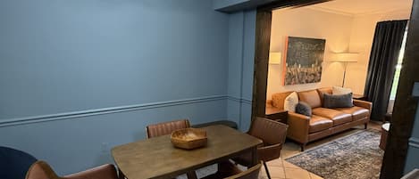 Un comedor con mesa y sillas de madera, una araña y vista a una sala de estar con sofá y lámpara.