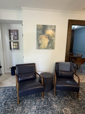 Ein Wohnzimmer mit zwei Ledersesseln, einem runden Hocker, einem gerahmten abstrakten Gemälde und einer kleinen Sammlung gerahmter Gegenstände an der Wand.