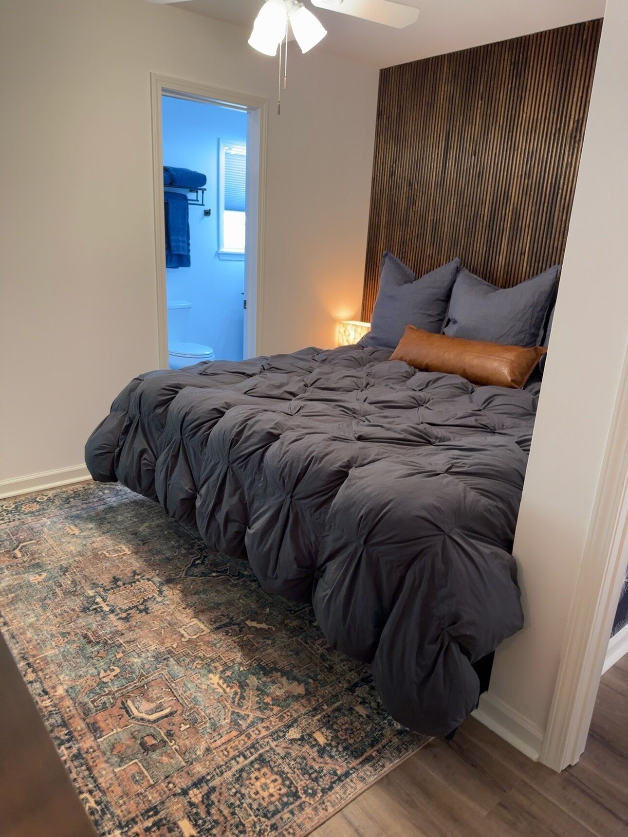 A bedroom with a large bed, a patterned rug, a ceiling fan, and a view into the bathroom.
