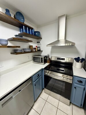 A compact kitchen with a dishwasher, microwave, stove, and oven. There are wooden shelves above the counter holding dishes and a blender. - Walk to Square or Campus from 2-bedroom / 2.5 bath condo in charming Oxford (Oxford)