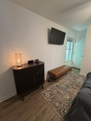 A modern living room with a wooden floor, a bench, a patterned rug, a wall-mounted TV, and a decorative lamp. - Walk to Square or Campus from 2-bedroom / 2.5 bath condo in charming Oxford (Oxford)