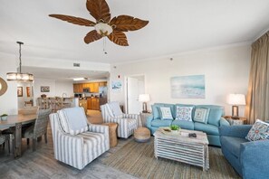 Een woonkamer met een plafondventilator, een blauwe bank, gestreepte fauteuils, een salontafel, en een eethoek met een houten tafel en stoelen.