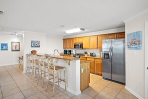 A kitchen with wooden cabinets, a stainless steel refrigerator, a microwave, and a bar stool area. - Snowbird Rates Hooked Bliss Bay Views Boat Slip (Gulf Shores)