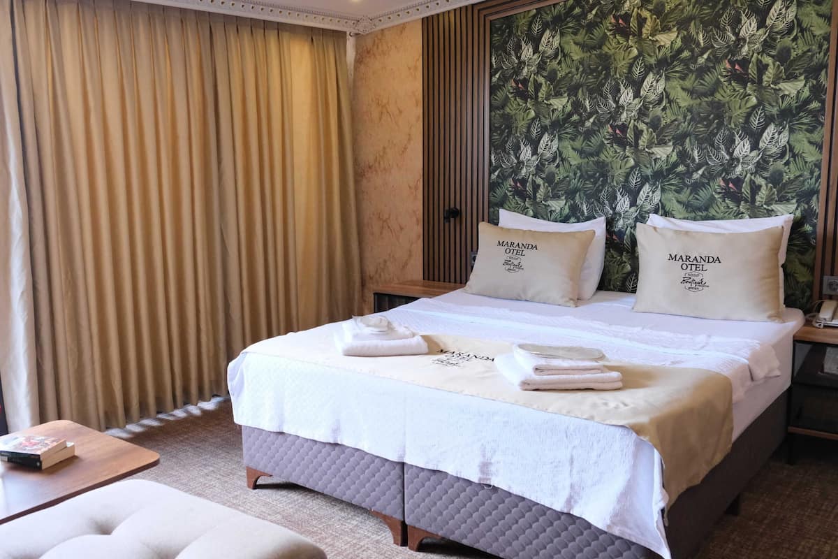 A hotel room with a bed, two pillows with 'MARANDA OTEI' branding, a bedside table with a phone, and a view of a wooden deck through a window.
