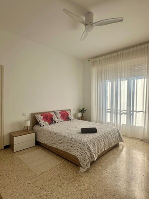 A bedroom with a bed, a nightstand, a ceiling fan, a window with sheer curtains, and a potted plant. - Moelda Home | Milan-Malpensa MXP 15′ Milan-Fiera (legnano)