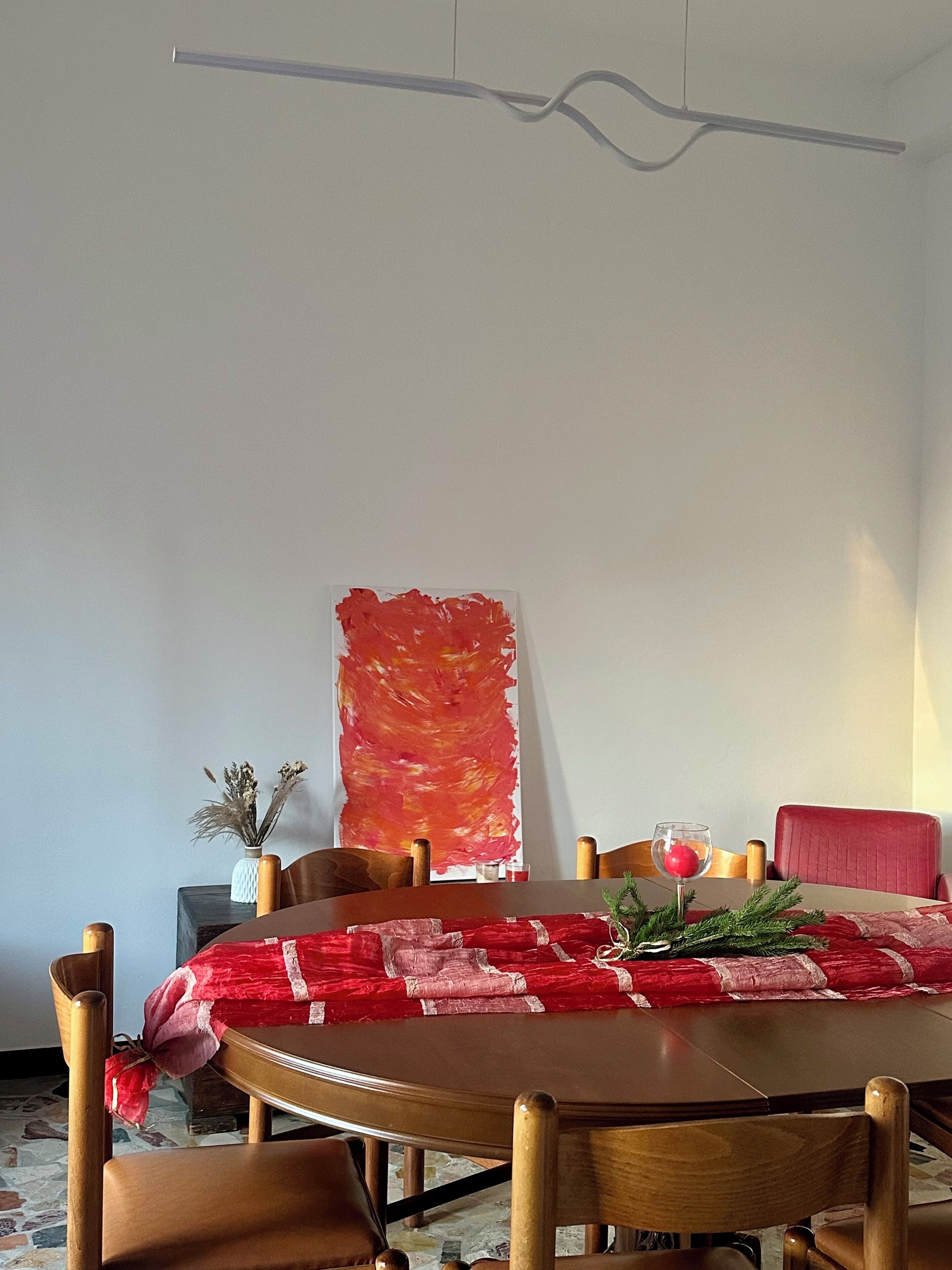 A dining area with a round table, wooden chairs, a red tablecloth, a red chair, a vase with dried flowers, and a large abstract painting on the wall.