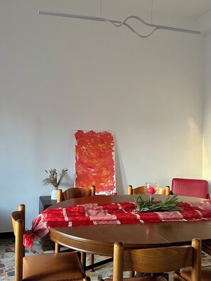 A dining area with a round table, wooden chairs, a red tablecloth, a red chair, a vase with dried flowers, and a large abstract painting on the wall. - Moelda Home | Milan-Malpensa MXP 15′ Milan-Fiera (legnano)