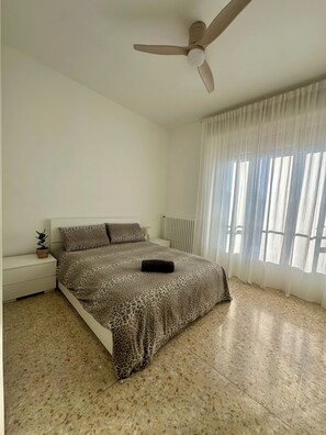 A bedroom with a bed, a ceiling fan, a radiator, a nightstand, a potted plant, and a window with sheer curtains. - Moelda Home | Milan-Malpensa MXP 15′ Milan-Fiera (legnano)