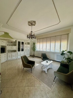 A spacious living room with a chandelier, two armchairs, a coffee table, and a large window with patterned curtains. - Moelda Home | Milan-Malpensa MXP 15′ Milan-Fiera (legnano)