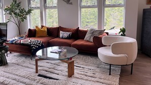 Een woonkamer met een bruine bank, een glazen salontafel, een witte fauteuil en een groot raam met uitzicht op bomen.