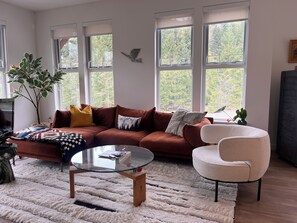 A living room with a brown sofa, a glass coffee table, a white armchair, and a large window with a view of trees. - Modern Mountain Retreat with Lake Views | 90+ stay (Britannia Beach)