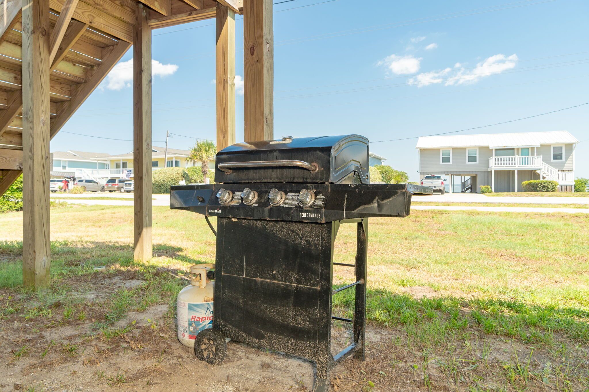 A black outdoor grill with a gas tank, set up under a wooden structure with a clear view of a residential area in the background.
