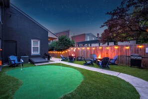 A backyard with a green artificial turf, a wooden fence, and outdoor seating with chairs and a barbecue grill. - The Stables | Walkable, MiniGolf, Fire Pits, Games (Louisville)
