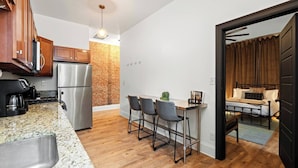 A compact kitchen with a refrigerator, a coffee maker, and a brick wall. There is a dining area with a table and chairs. - The Stables | Walkable, MiniGolf, Fire Pits, Games (Louisville)