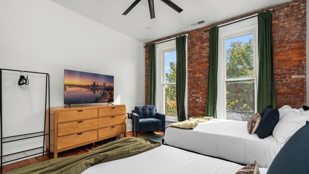 A bedroom with a bed, a wooden dresser, a chair, and a large window with curtains. - The Stables | Walkable, MiniGolf, Fire Pits, Games (Louisville)
