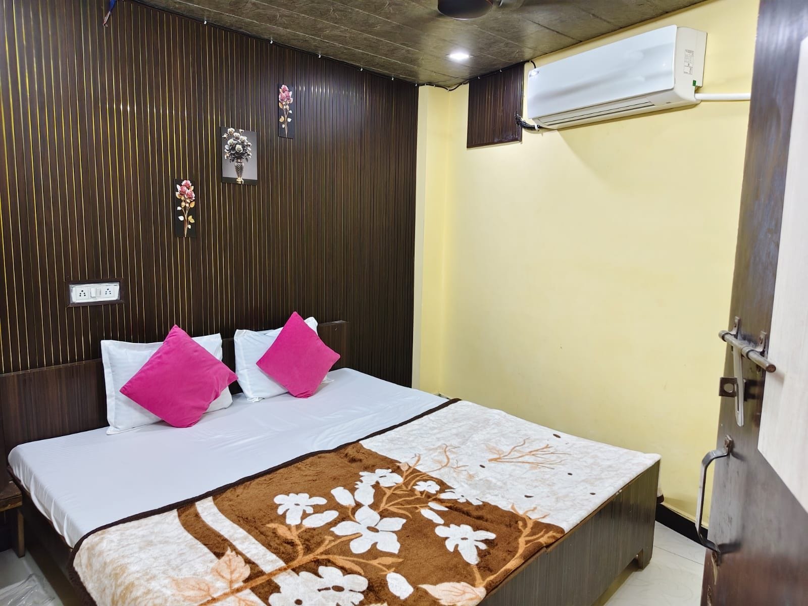 A hotel room with a bed, a wall-mounted air conditioner, and a power outlet.