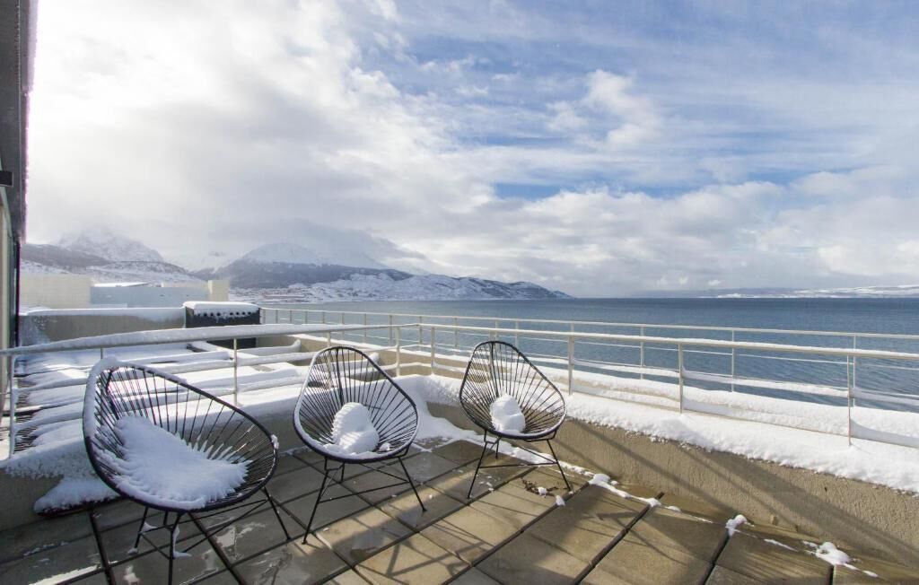 A deck with three chairs and a table, overlooking a snowy landscape and a body of water.