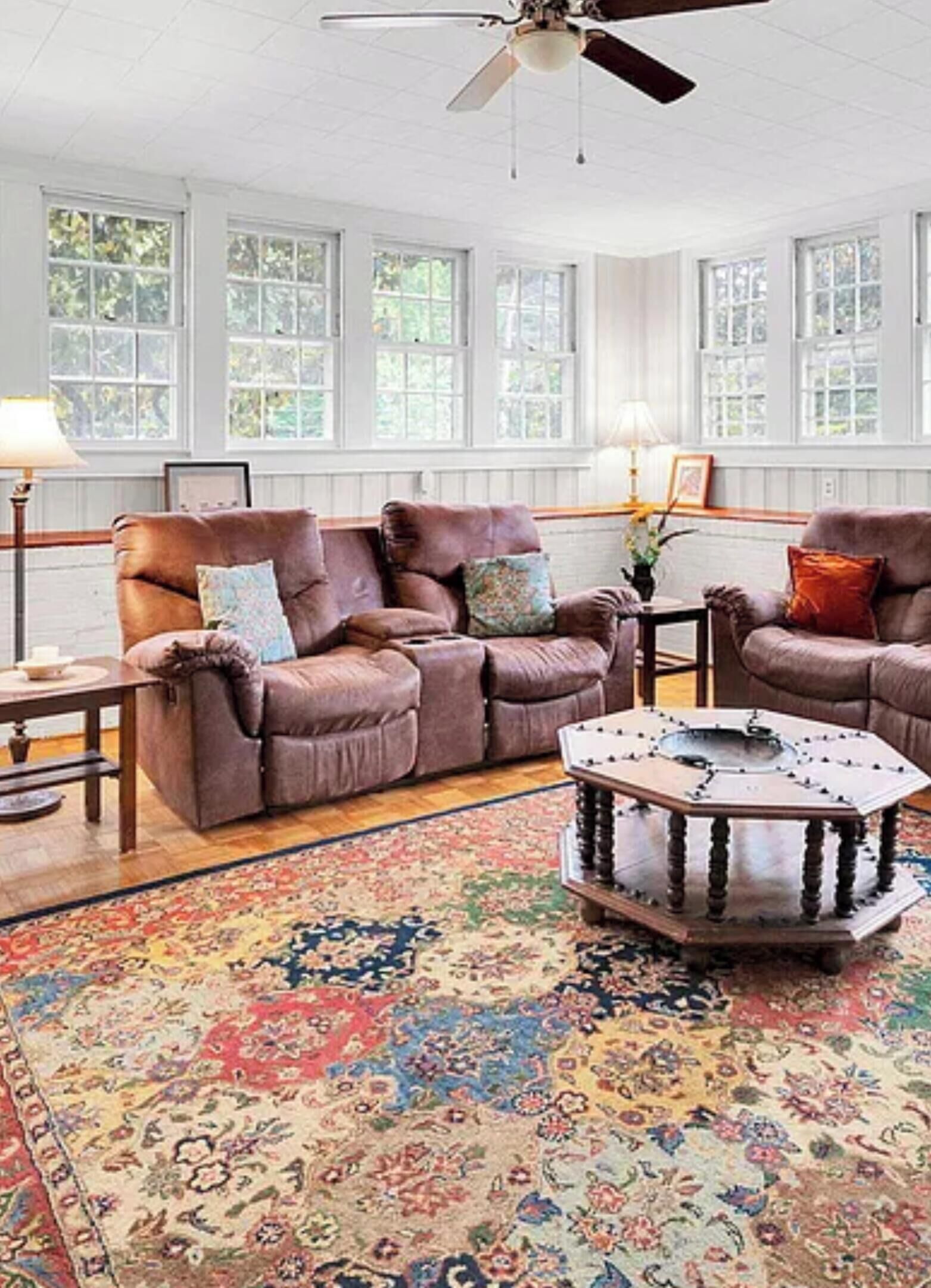 Ein geräumiges Wohnzimmer mit Deckenventilator, großen Fenstern und einem gemusterten Teppich.