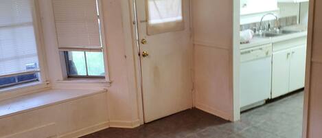 A room with a window seat, a door with a window, and a kitchen area with cabinets and a sink.