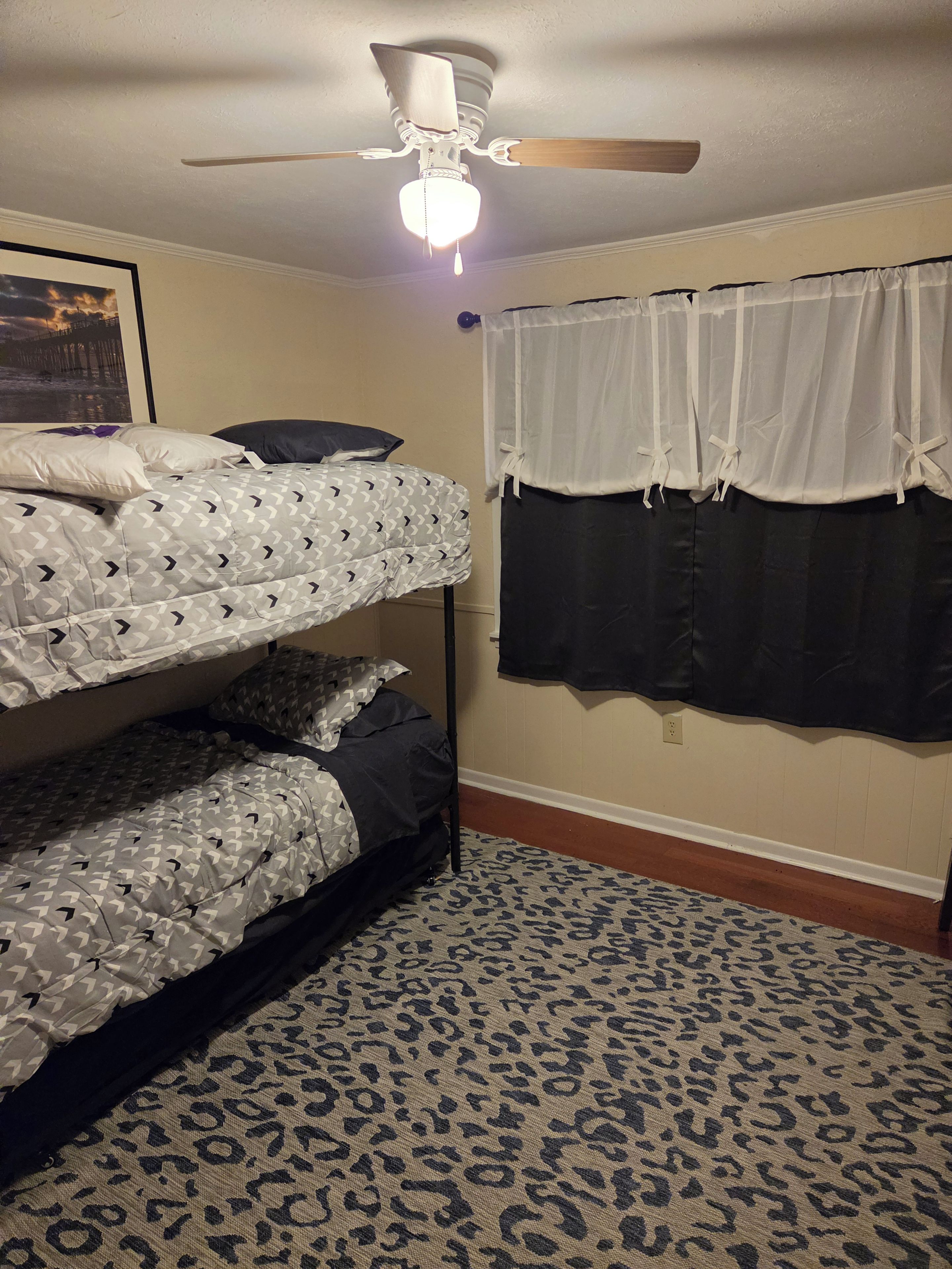 A room with a bunk bed, a ceiling fan, a window with curtains, and a patterned rug.