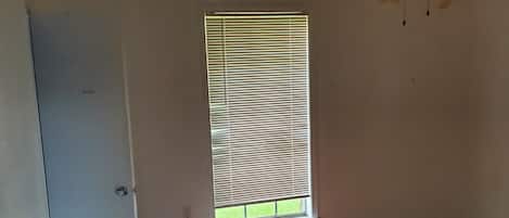 A room with a wooden floor, a window with blinds, a ceiling fan, and a white door.