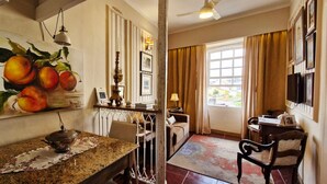 A hotel room with a dining area, a painting of fruit on the wall, a chandelier, and a view of the outside. - COPEL0100 - Prédio 34 (Salvador)