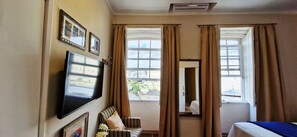 A hotel room with a bed, a television mounted on the wall, a window with curtains, and a view of the outside. - COPEL0100 - Prédio 34 (Salvador)