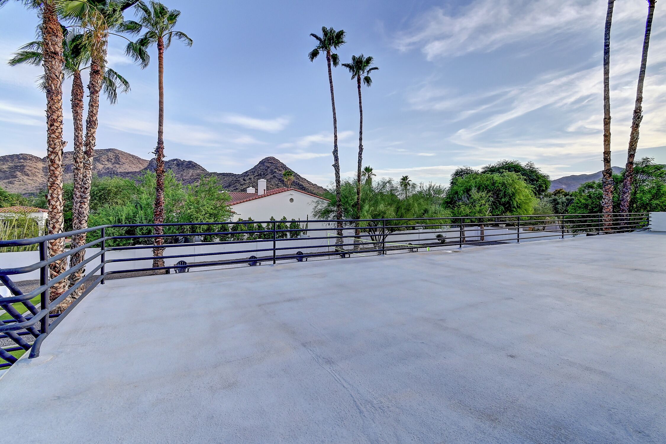 An outdoor terrace with a concrete floor, surrounded by palm trees and a mountain in the background.