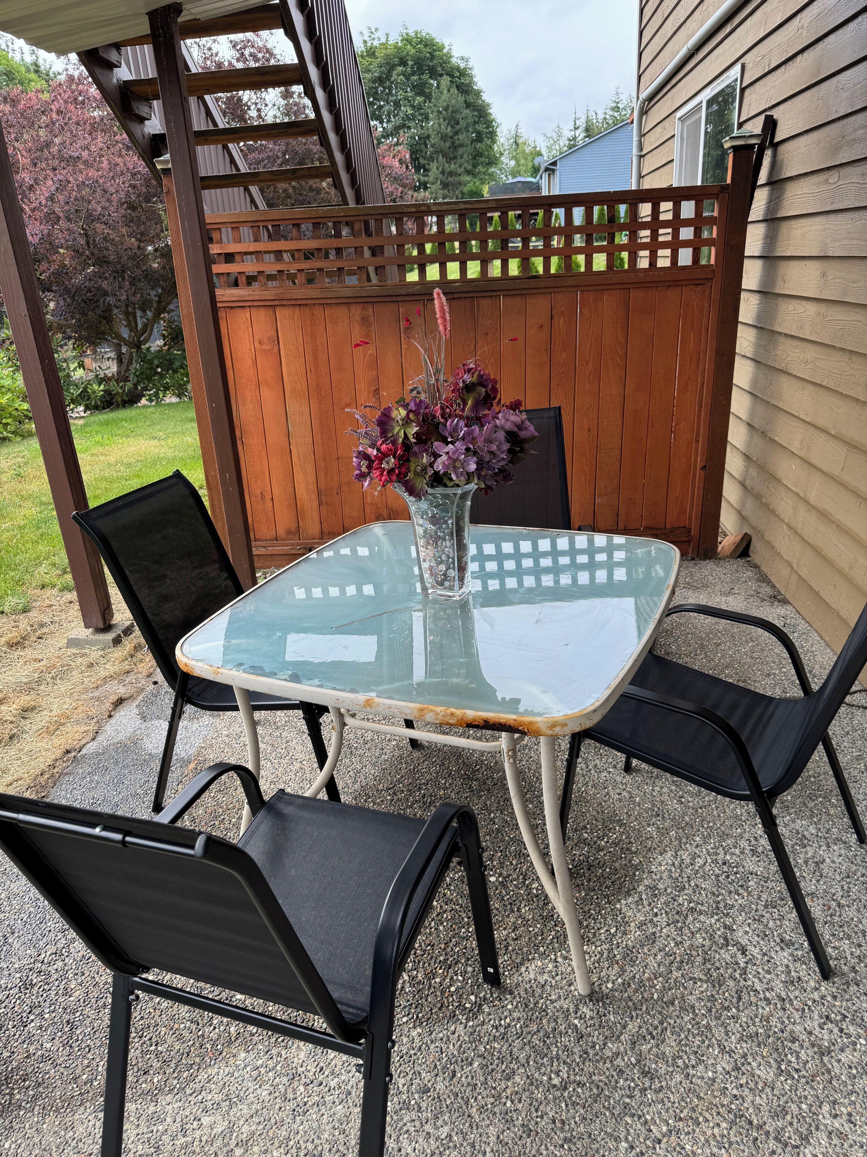 Une terrasse extérieure avec une table en verre et quatre chaises, une clôture en bois et une maison en arrière-plan.