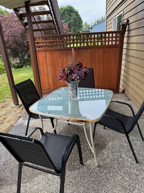 Eine Terrasse im Freien mit einem Glastisch und vier Stühlen, einem Holzzaun und einem Haus im Hintergrund.