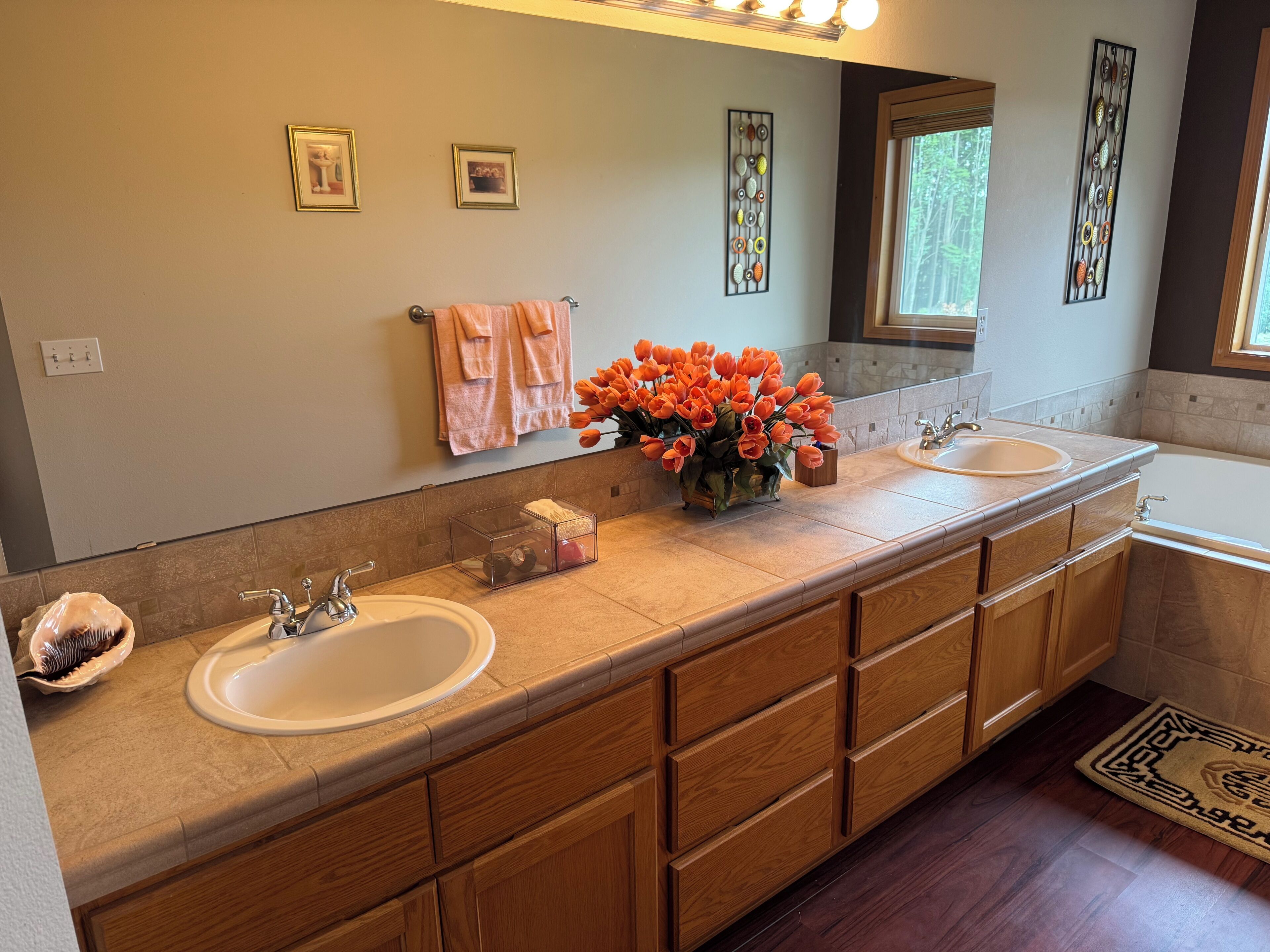 Une salle de bain moderne avec deux vasques, des meubles en bois, un grand miroir et un vase décoratif rempli de fleurs oranges.
