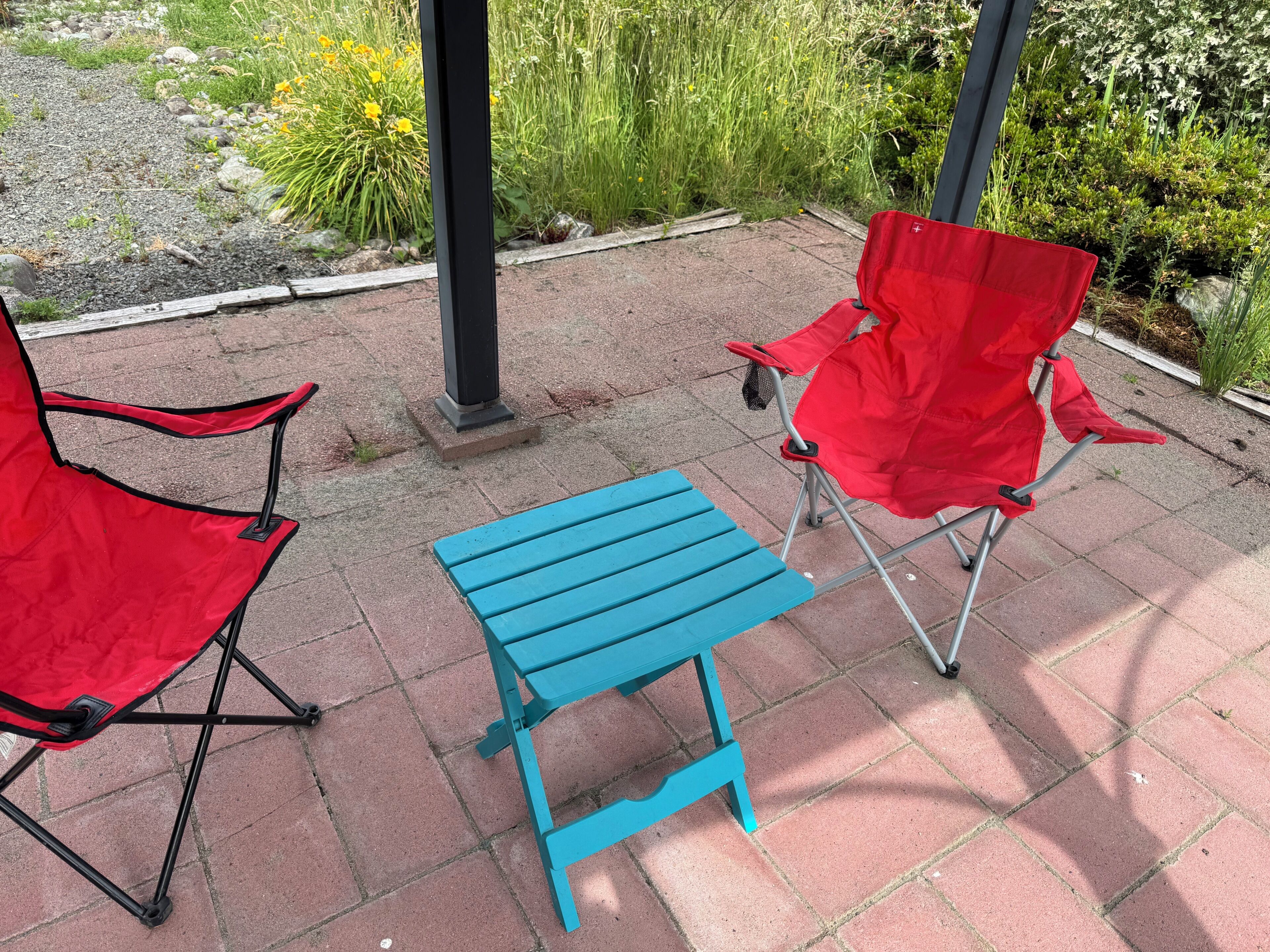 Zwei rote Klappstühle und ein blauer Tisch auf einer gepflasterten Terrasse mit Garten im Hintergrund.