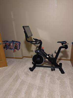 Een stilstaande hometrainer met een digitaal display, geplaatst in een kamer met vloerbedekking en een deur links.