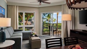 A hotel room with a sofa, ottoman, dining table, chairs, a TV, and a balcony with a view of palm trees and a building. - ALOHA!! GRAND OCEAN VIEW  1-bedroom Villa with AC in Kaanapal, Steps to Beach! (Lahaina)